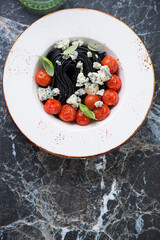 Plate with black spaghetti, blue cheese, roasted tomatoes and green basil, above view on a black marble background, vertical shot with space