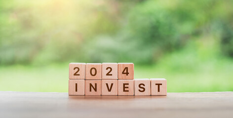 wooden blocks spelling ‘2024 INVEST’ on a rustic table, set against a blurred backdrop of lush...