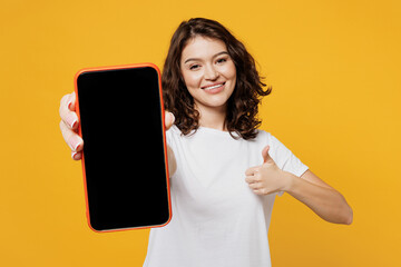 Obraz premium Young woman wear white blank t-shirt casual clothes hold in hand use blank screen workspace area mobile cell phone show thumb up isolated on plain yellow orange background studio. Lifestyle concept.