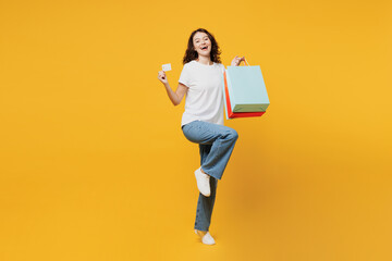 Full body young woman wears t-shirt casual clothes hold in hand paper package bags after shopping show credit bank card isolated on plain yellow orange background. Black Friday sale buy day concept. - obrazy, fototapety, plakaty
