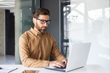 Serious concentrated thinking businessman working inside office with laptop, mature enlightened man in shirt and glasses at workplace typing on keyboard, programmer developer