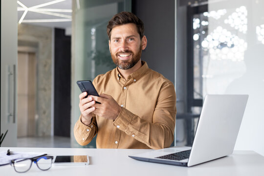 Portrait of mature successful businessman inside office, male looking at camera smiling, worker in shirt holding phone, using online application