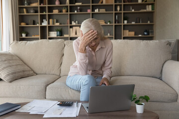 Unhappy old woman sit on sofa in living room, manage finances, suffer from lack of money, calculates expenses using laptop and calculator, feeling anxious about debt or bankruptcy, financial crisis