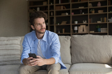 Serious thoughtful young man 35s sitting on sofa with cellphone in hands, deep in thoughts looking...