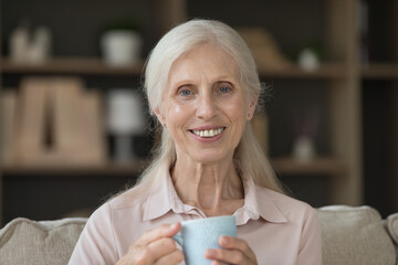 Close up head shot portrait of happy attractive senior single woman spend free time sitting on sofa, smiling, posing, look at camera, holds cup of tea, enjoy hot beverage and quiet life on retirement