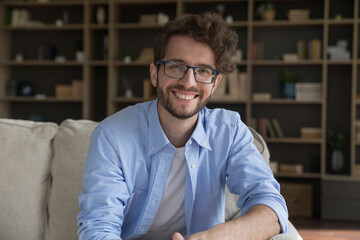 Happy millennial 35s handsome man sit on sofa posing, staring at camera. Bearded guy in eyeglasses spend time at home, enjoy carefree relaxation at own or rented apartment. Leisure, homeowner portrait