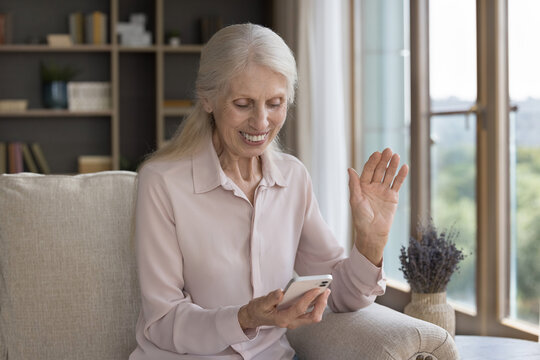 Positive mature female sit on sofa look at cellphone screen enjoy good on-line news, read notice with sell-out commercial offer. Elderly woman start videocall to family use videocall app on smartphone