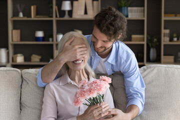 Millennial caring son covering mature mothers eyes with palm, gives her carnation flowers, makes pleasant surprise during his visit at mommy home. 8-march or life event celebration, attention, love