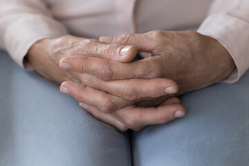 Close up of old woman hands folded on laps sitting indoors, feels lonely, desperate or anxious at...