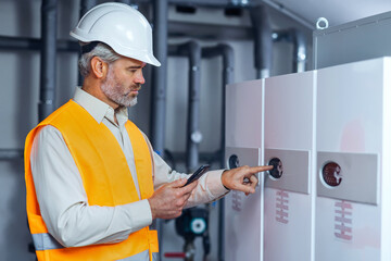 Professional Industry Engineer Worker Wearing Safety Uniform and Hard Hat Uses phone for controlling equipment. Serious Successfulmale Industrial Specialist in a boiler room