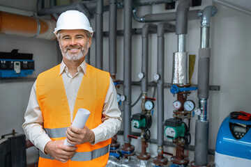 senior gray haired Dedicated smiling factory worker standing next to pipes with gas pressure. Worker is dressed in protective uniform, having hardhat