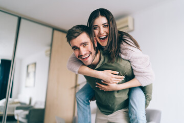 Happy man giving piggyback ride to laughing girlfriend in apartment