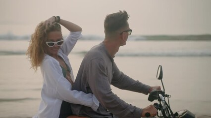 Couple in sunglasses ride motorcycle on sea beach at sunset. Lady in swimwear hugs guy, touches wind blown hair, smiles at camera on romantic bike drive. Man, woman travel on motorcycle by ocean.