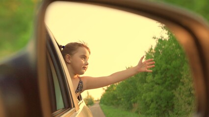 Child smiles, enjoys family vacation, car trip. Happy child is driving in car, looking out window with an outstretched hand. Little girl, child traveling by car, Video in rear view mirror. Child trip - obrazy, fototapety, plakaty