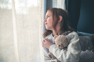 a pensive cute girl sits and looks out the window, hugging a teddy bear.