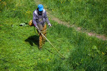 lawnmower man with a weed eater or a string trimmer trimming grass at sunny day.