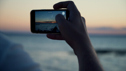 Young Man Capturing a Stunning Sunset on the Seashore with His Smartphone, Focusing on the Close-Up of the Phone in His Hands, Showcasing the Beauty of Nature and Photography.