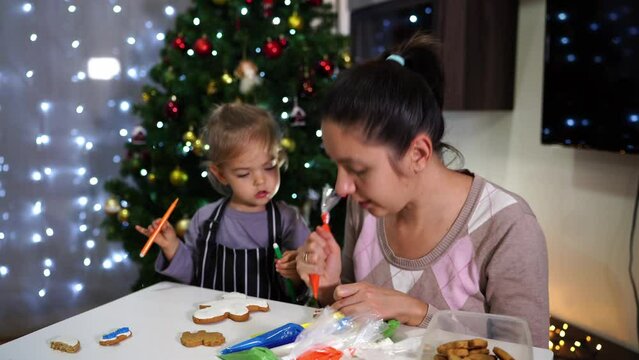 Little girl stands next to her mother painting icing on cookies at the table
