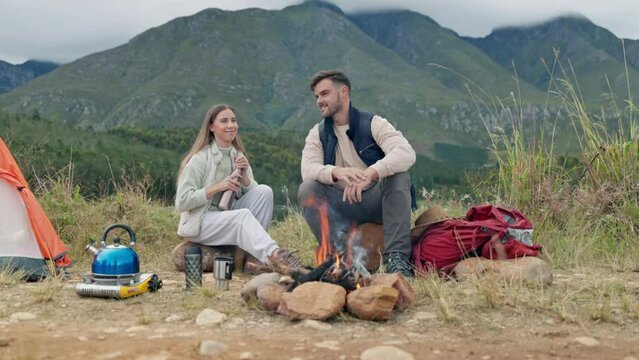 Couple, camping and fire with wood, coffee and relax in mountains, nature or forrest for holiday. Man, woman and campfire on ground for cooking, giving thermos and vacation in bush, field or morning