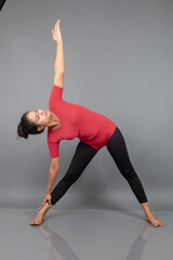 Healthy young woman in red t-shirt and black leggings doing yoga and stretching exercises isolated on grey background