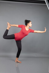 Healthy young woman in red t-shirt and black leggings doing yoga and stretching exercises isolated on grey background