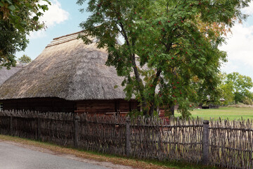 An ancient traditional fence made of wicker, an ancient farmhouse with a thatched roof behind it. View of the courtyard of an old Lithuanian village