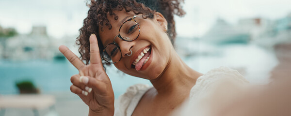Closeup of a young woman in glasses stands in the seaport and shots selfie on smartphone. Positive woman using mobile phone outdoors in urban background. - obrazy, fototapety, plakaty