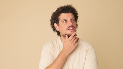 Thoughtful guy with curly hair dressed in beige t-shirt makes choice, ponders an idea while...