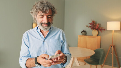 Happy elderly man with gray hair wearing blue shirt while standing in the living room in modern apartment exchanging text messages