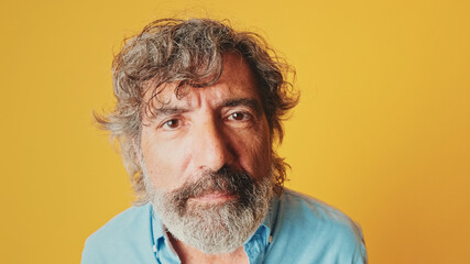 Close-up, of an elderly grey-haired bearded man wearing a blue shirt, looking at the camera trying...