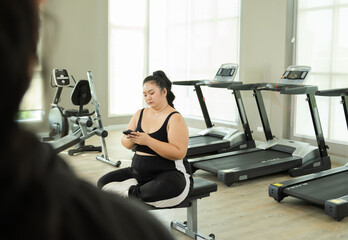 Happy Asian overweight woman use smartphone looking video online relaxing in fitness club, chubby...