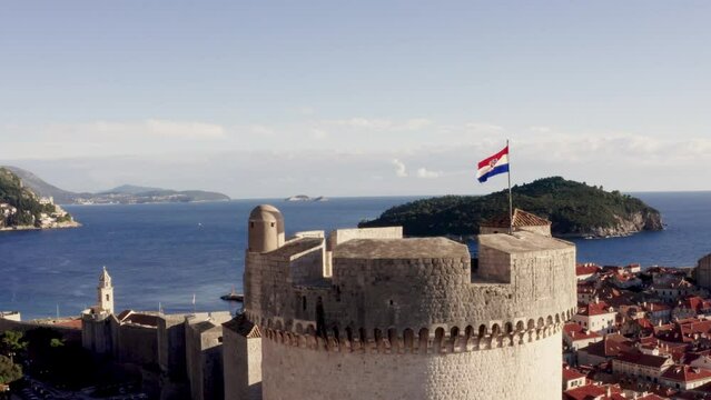 Drone flies over historic old town Dubrovnik Croatia capturing a beautiful aerial shot of castle wall lookout tower with flapping flag in the sunlight over the Adriatic Sea 