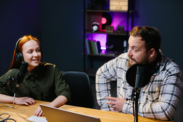 two radio hosts in headphones laughing while recording podcast in studio together.