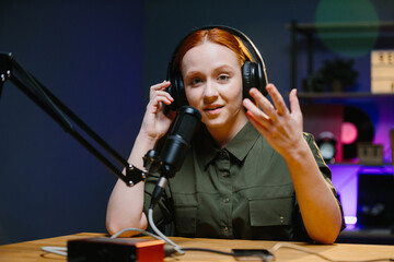 Modern young adult woman wearing headphones recording podcast or broadcast in home studio.