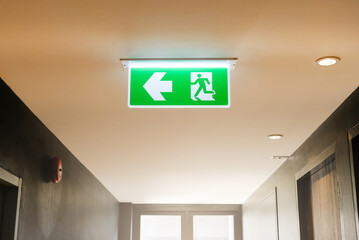Lighted glowing green emergency exit signs in an office hallway with arrows pointing the way out of...