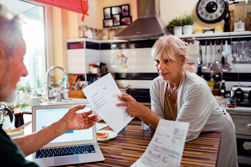 Senior couple doing home financials in kitchen. Mature woman holding phone bill showing to husband