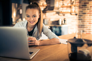 Smiling young woman with coffee cup sitting at desk with laptop