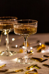 party, celebration and holidays concept - close up of champagne in glasses, electric garland lights and golden confetti on table over black background