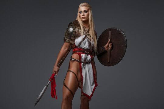 Viking woman, clad in fantasy armor with bare thighs. She confidently wields a shield and spear on a gray backdrop