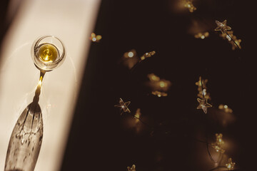 One glass of cognac sparkling at sunlight, dark background with star garlands, shadow, sun flare light on table. Strong drink with star filter, top view, muted colors, winter holidays