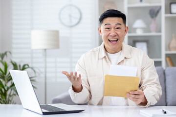 Surprised Asian man sitting at desk in home office, holding open envelope in hands, can't believe...
