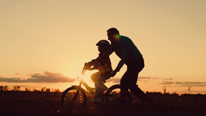 Father helps his daughter ride bike. Father teaches child wearing safety helmet to ride bicycle in park. Child rides bicycle. Kid, dad play together, sunset. Child dream learns to ride bicycle. Family - obrazy, fototapety, plakaty