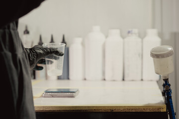 A woman in black protective gloves puts a plastic cup on the scales in the workshop. An airbrush is hanging nearby. Bottles with the necessary materials are in the background