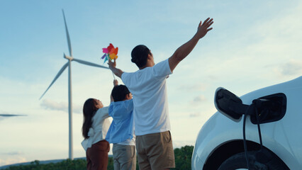 Concept of progressive happy family enjoying their time at wind farm with electric vehicle. Electric vehicle driven by clean renewable energy from wind turbine generator for charging station. - obrazy, fototapety, plakaty