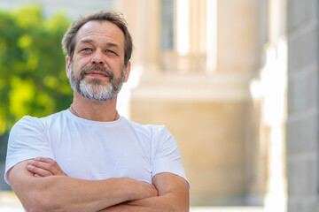 A handsome middle-aged man with a beard stands on a blurred city background, crossing his arms and smiling in a relaxed and joyful manner. success and optimism.