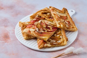 Sliced open apple pie on puff pastry with apple slices, sprinkled with almond petals, on a white ceramic board on a pink marble background.
