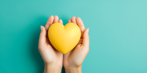 The woman hand is holding a yellow heart on teal blue background - obrazy, fototapety, plakaty