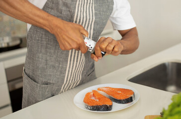 Unrecognizable black man seasons salmon with pepper in kitchen