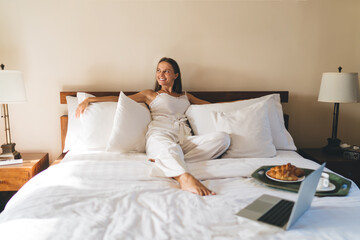 Cheerful woman resting with laptop and breakfast tray on bed - Powered by Adobe