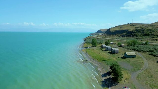An aerial view of a large blue azure sea lake in clear sunny weather. On the shore houses and boats, in the distance high mountains in the clouds. Drone video, flying in an arc. Lake Sevan, Armenia.